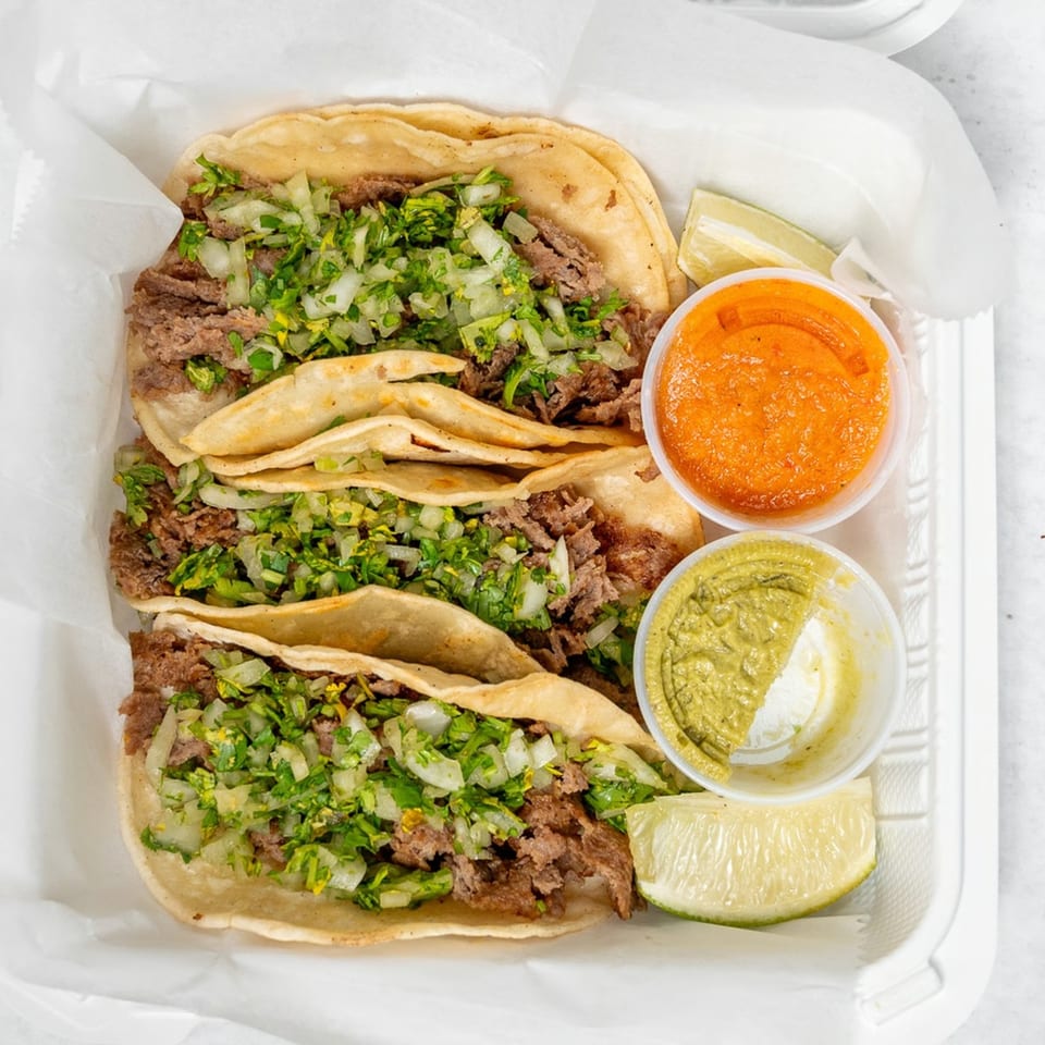 Three carne asada tacos with cilantro and onions, with red and green salsa and lime wedges.